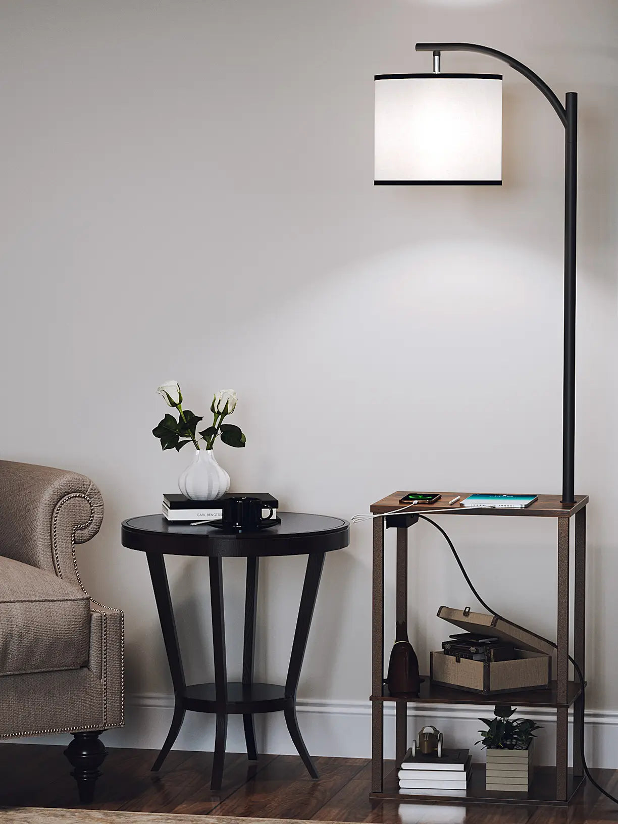An elegant arc floor lamp with a white drum shade and an integrated two-tier side table for display and charging, enhancing a living room seating area with a classic armchair and a round accent table.