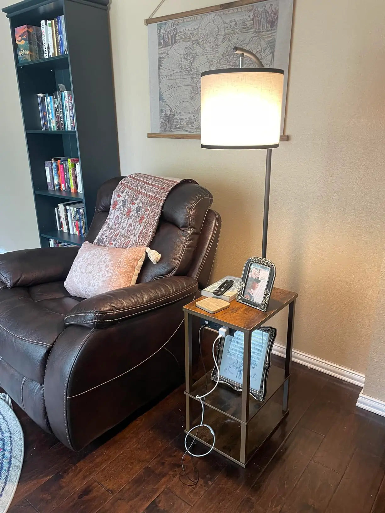 A rustic-style floor lamp with an integrated two-tier side table, providing warm light next to a comfortable brown leather recliner in a reading nook, with a bookshelf and decorative map
