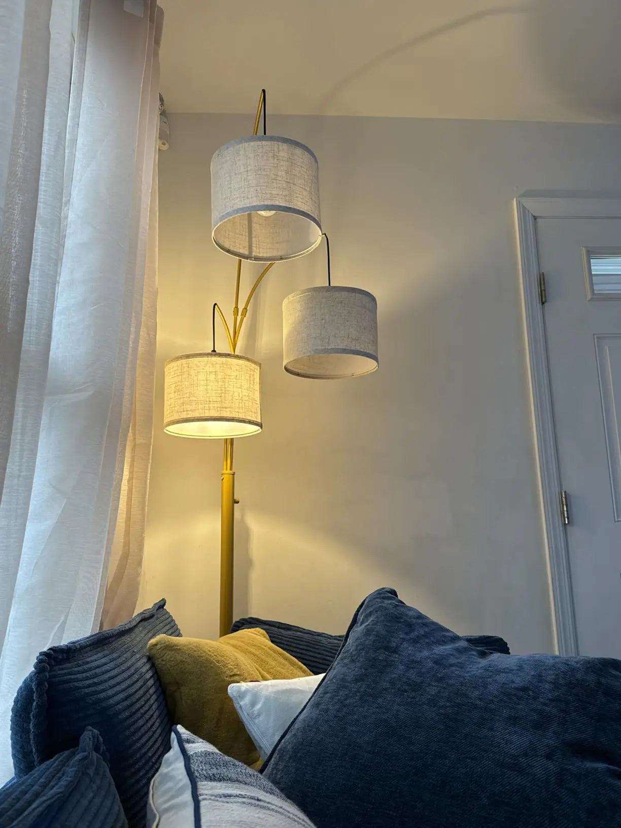 A transitional style 3-light arc lamp with neutral fabric shades positioned next to a window seat in a living room, providing warm light for a reading nook near a brown recliner.