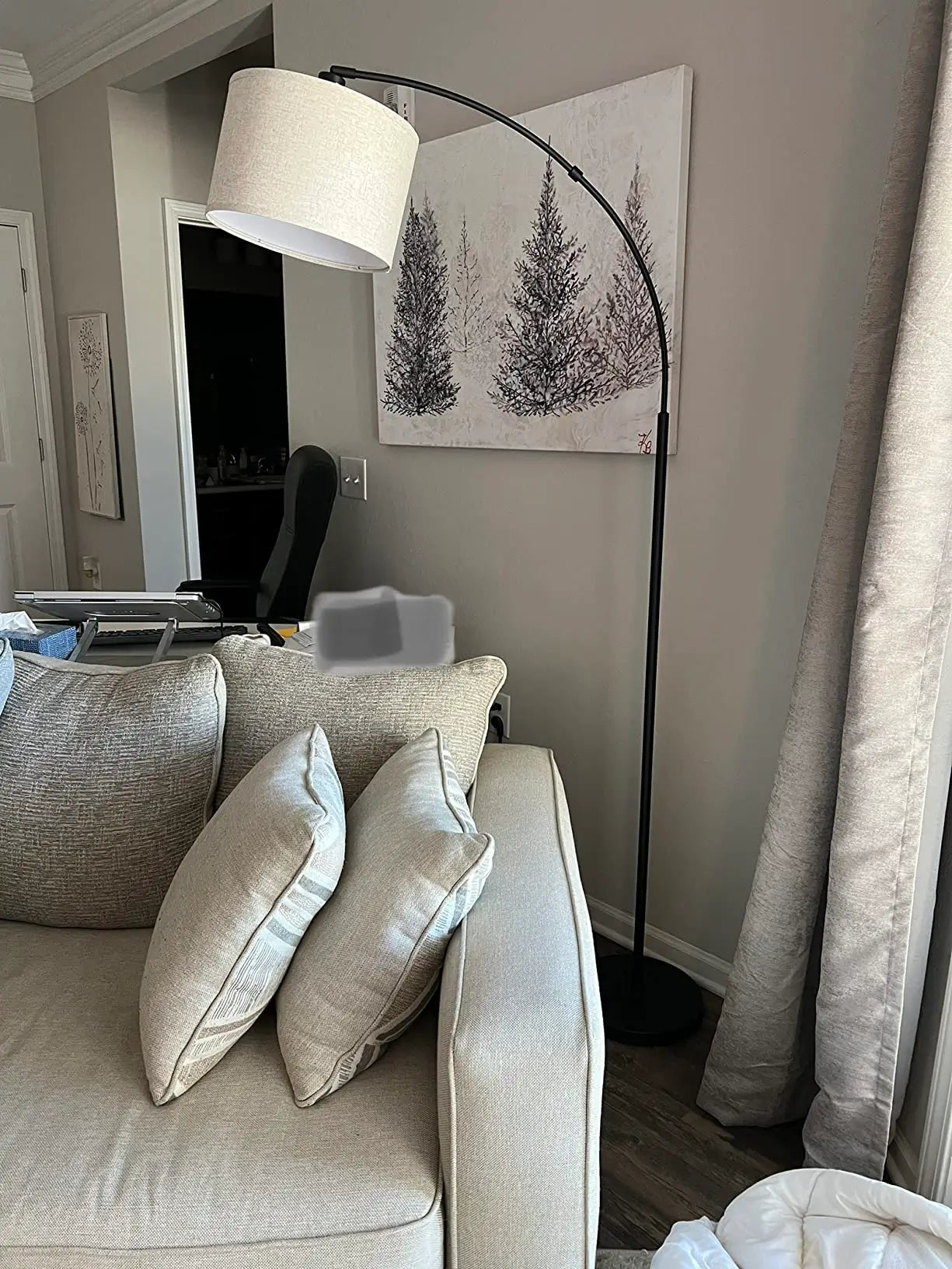 A contemporary arc floor lamp with a slender metallic frame and a large, neutral-toned fabric drum shade, gracefully extending over a comfortable grey sofa in a minimalist living room.