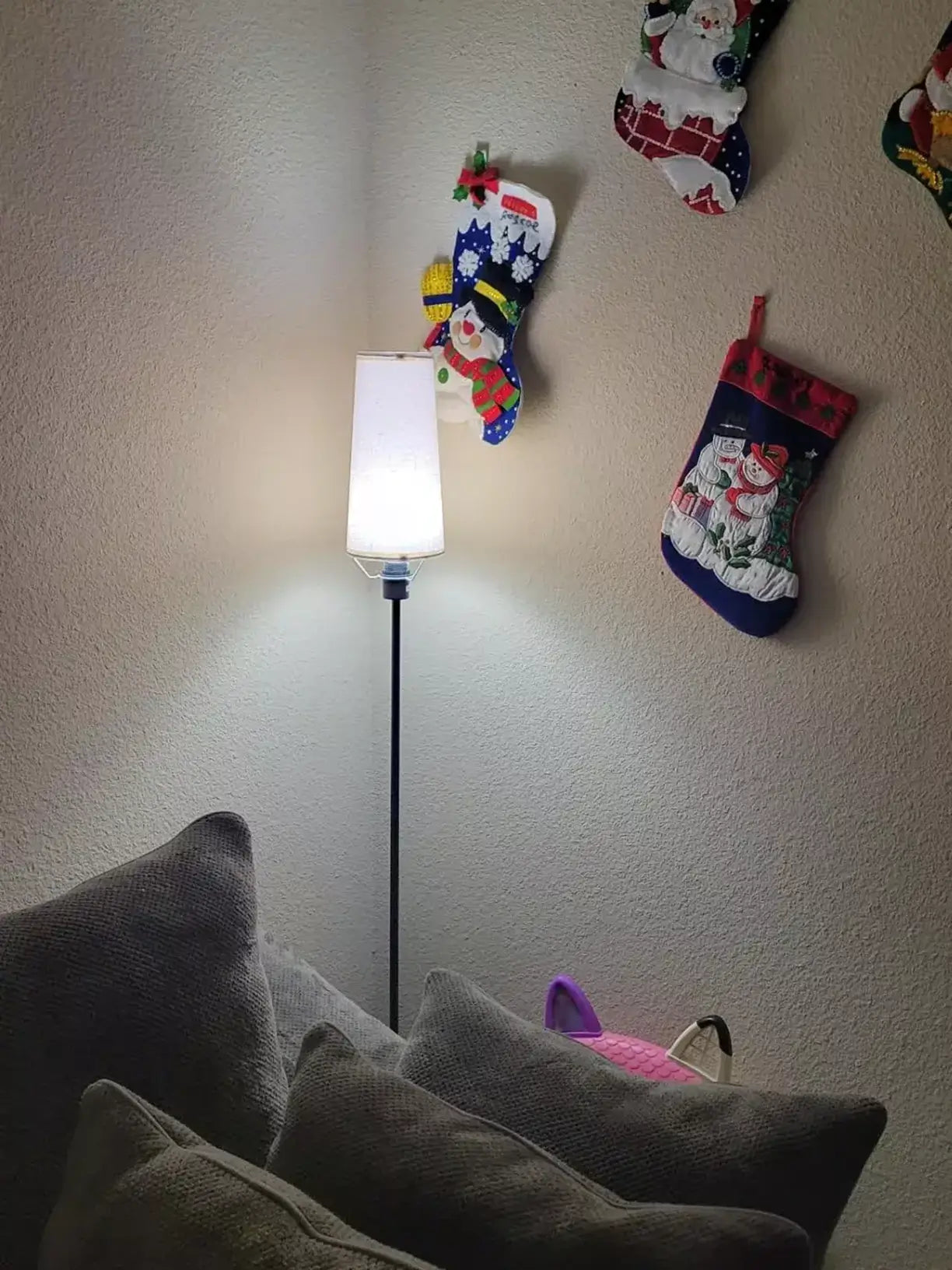 A minimalist floor lamp with a white cylindrical shade providing warm light in a room decorated with festive Christmas stockings hanging on the wall, with a grey couch in the foreground.