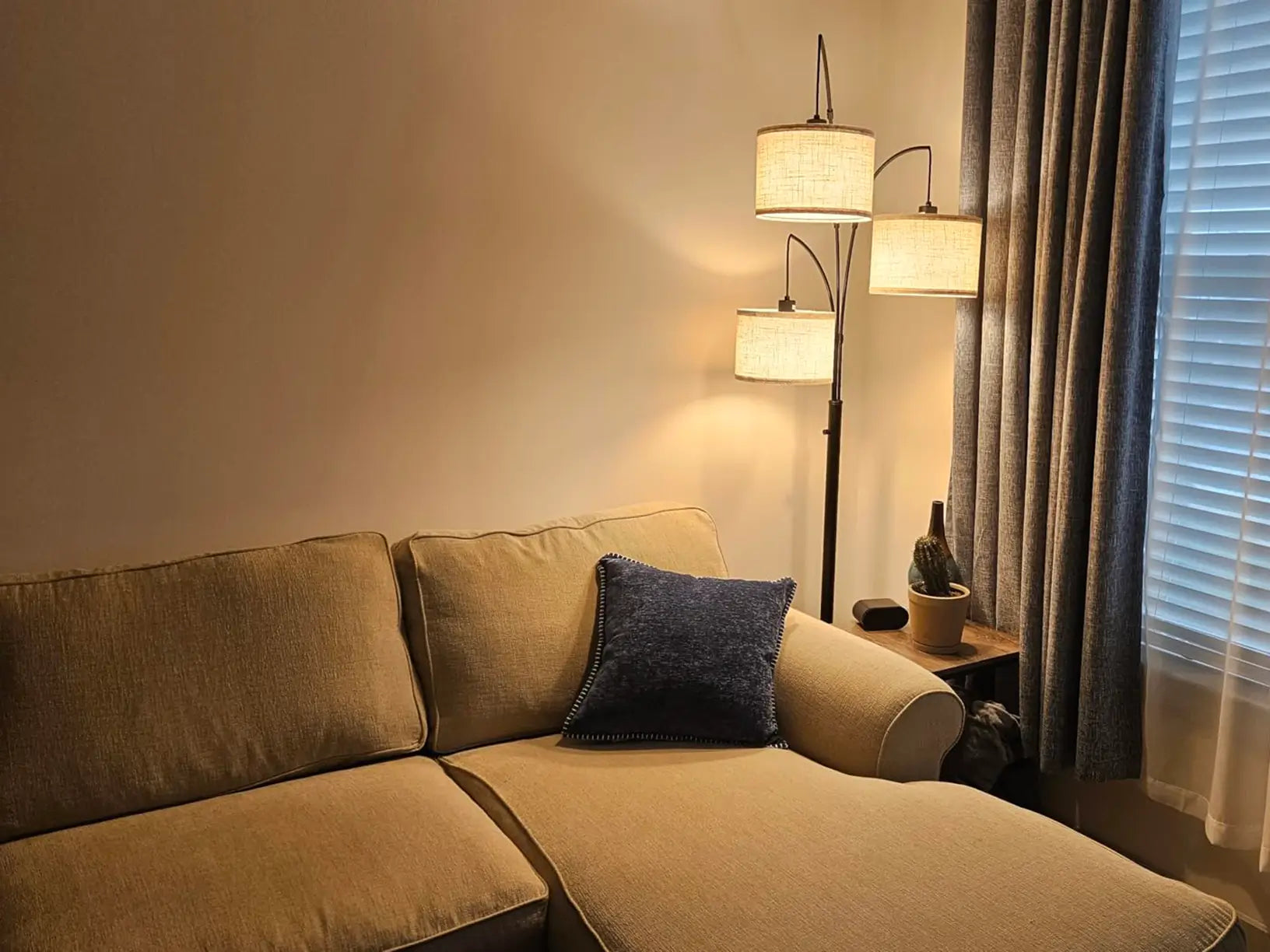 A minimalist triple-head arc floor lamp with light cylindrical shades standing behind a comfortable sofa with multiple pillows, providing ambient lighting in a modern interior.