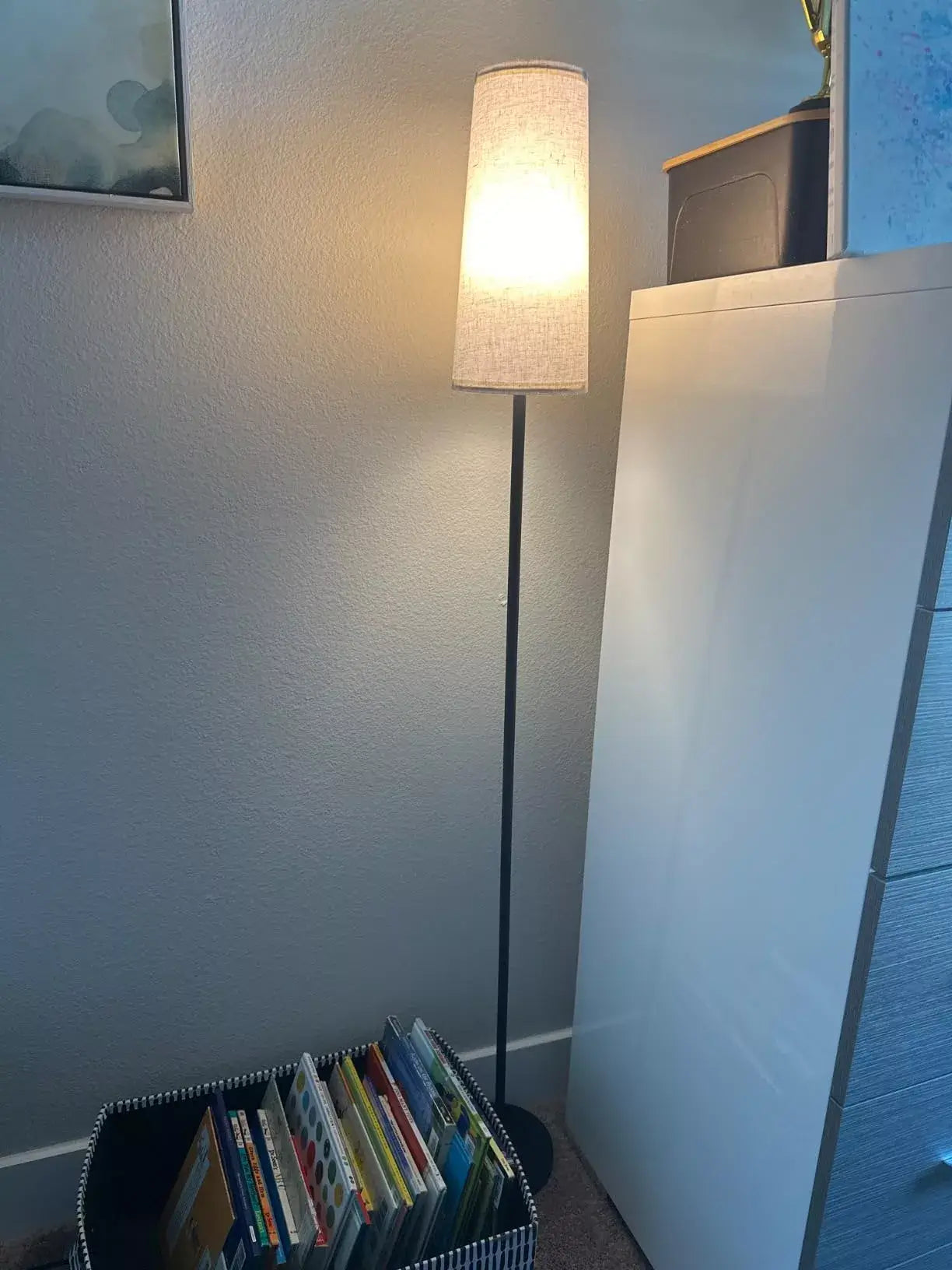 A tall, slender modern floor lamp with a light fabric shade illuminating a cozy reading corner in a living room, next to a white dresser and a basket of books.