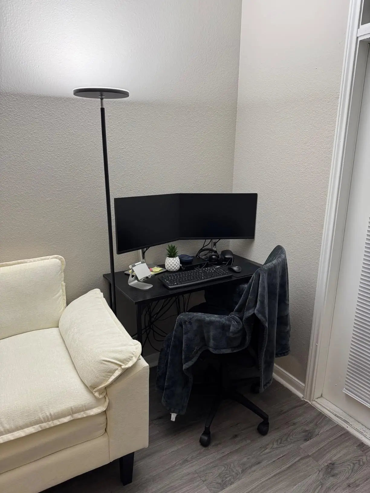 A slim LED torchiere floor lamp with a small, upward-facing light head, providing efficient and space-saving illumination in a modern home office or study area with a desk and monitor.