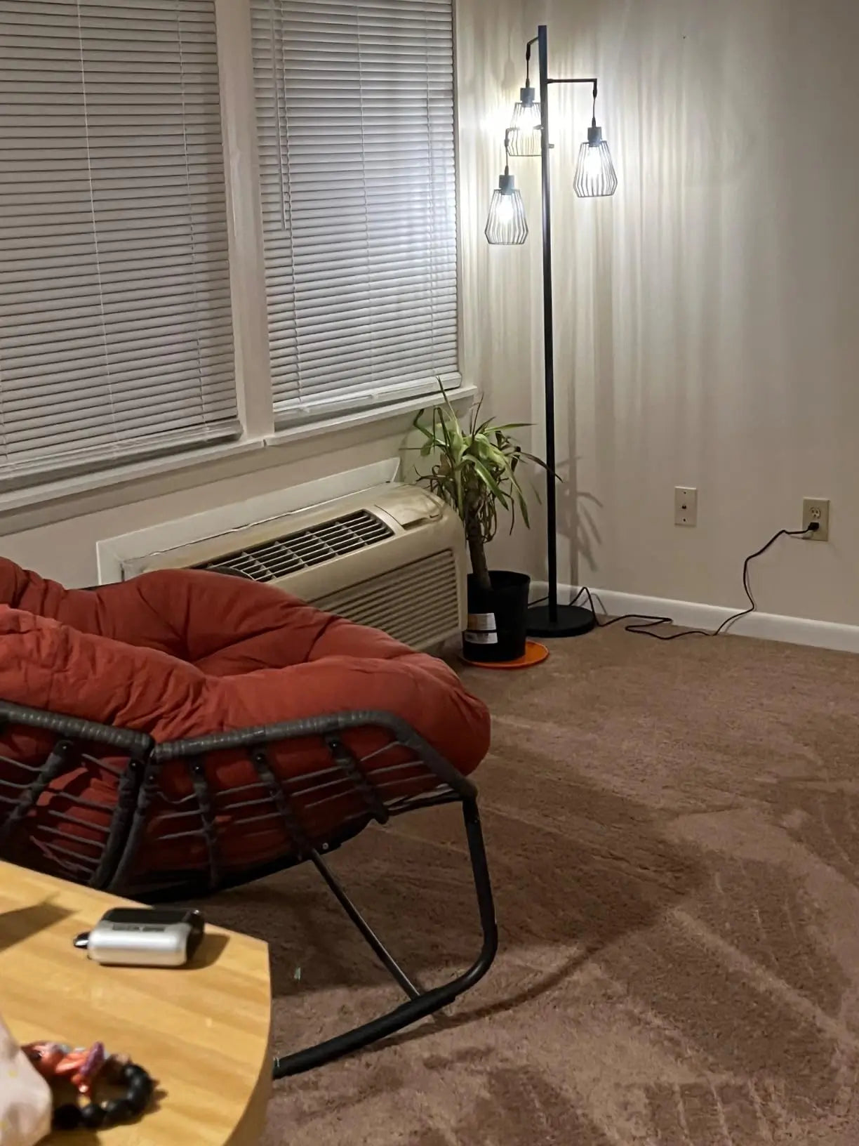 A tall black tree floor lamp with three industrial-style metal cage shades, casting warm light in a contemporary room corner with a large houseplant and a simple wall.